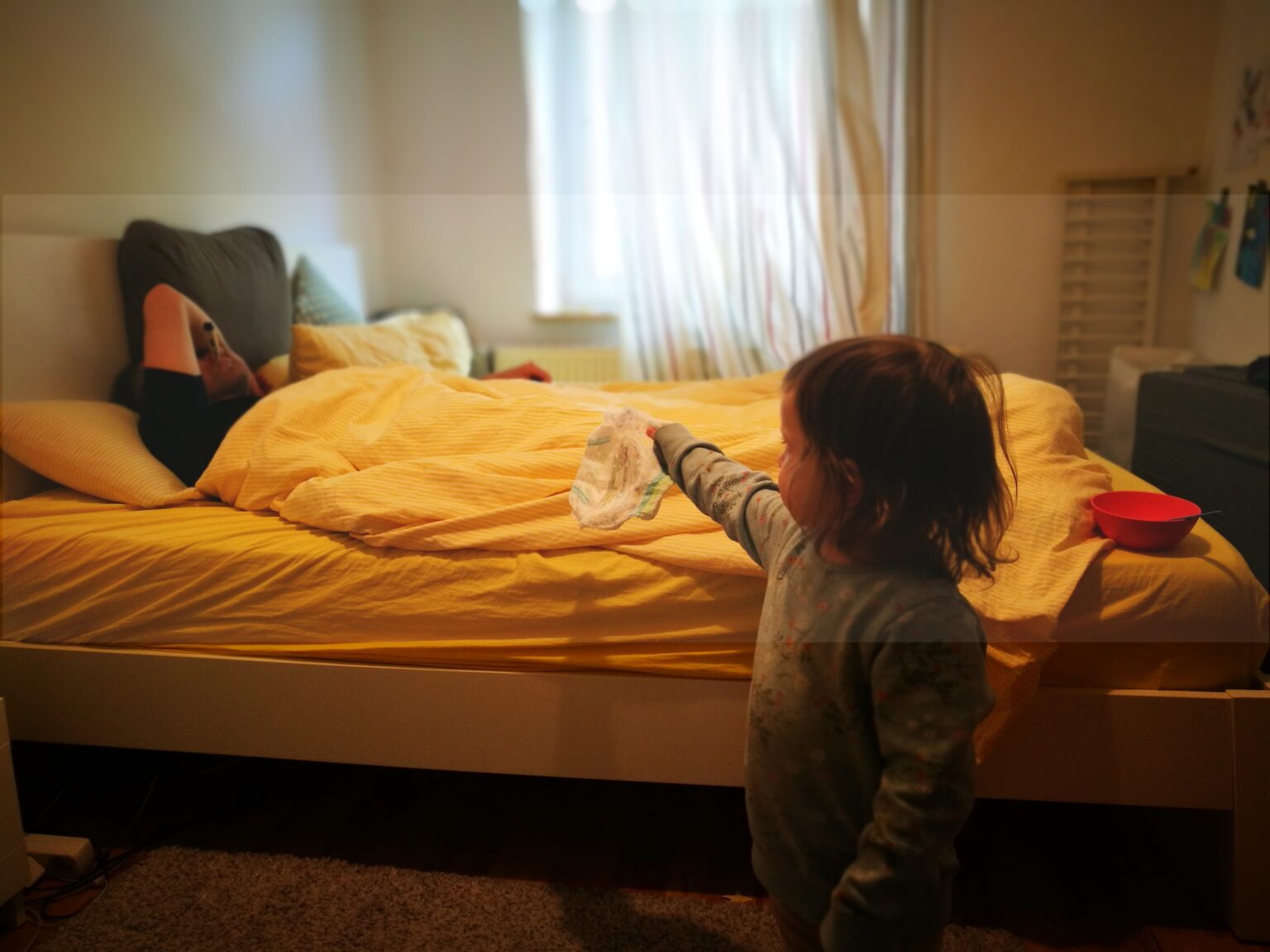Ein Kleinkind steht mit einer Windel in der Hand vor dem Bett des kranken Vaters