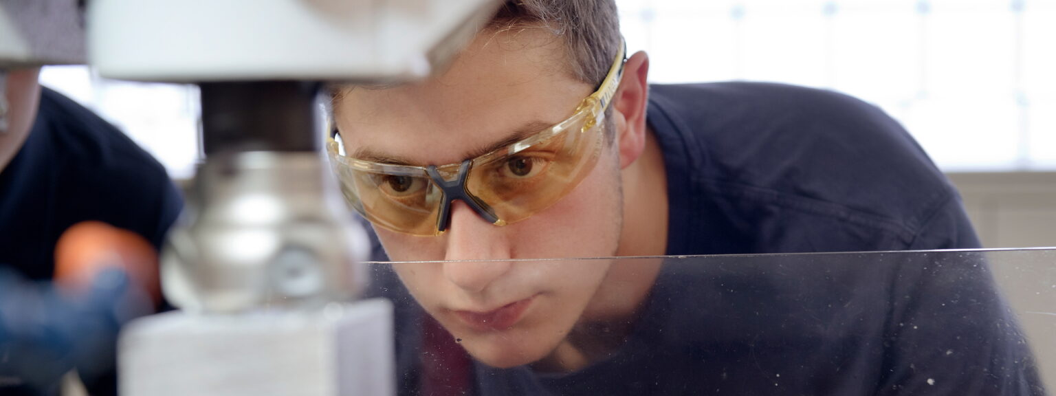 Ein junger Mann mit Schutzbrille schaut auf ein Werkstück in einer Maschine