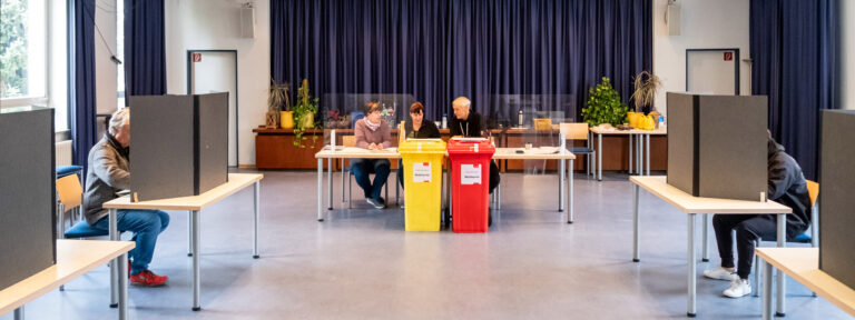 Landtagswahl 2023: Im Wahllokal in der Ingoldstädter Straße sitzen die Wahlhelfer:innen zwischen den gelb und roten Wahltonnen. Sie nehmen die Stimmzettel der Wähler:innen entgegen.