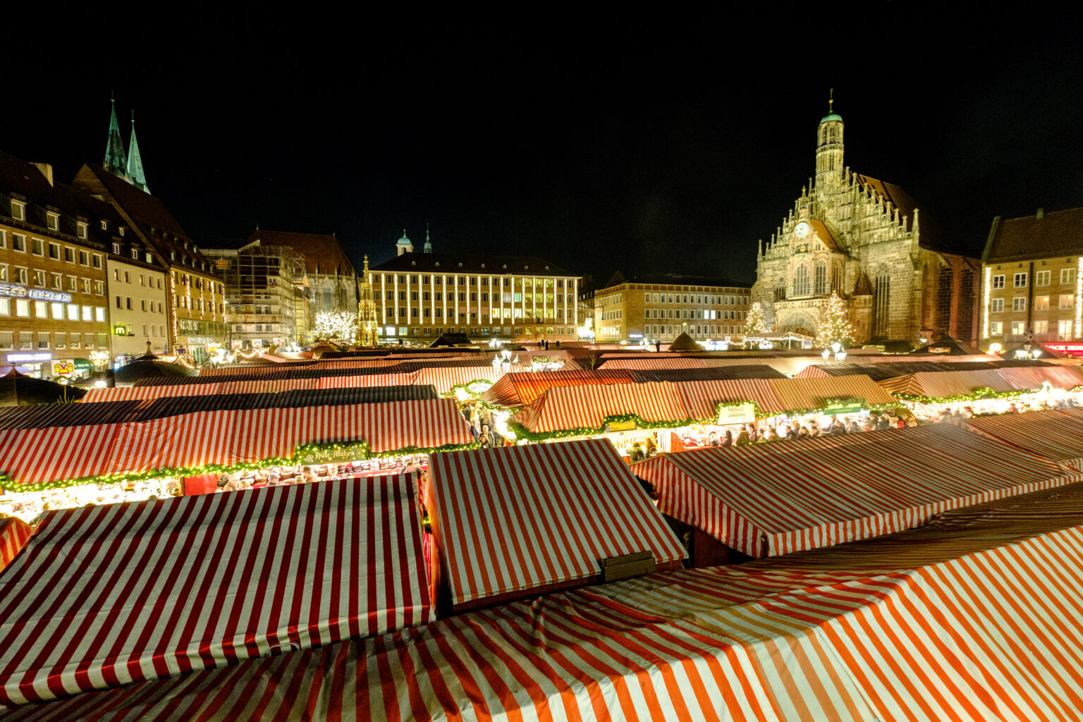 Ein Blick über den Nürnberger Christkindlesmarkt bei nacht. Im Vordergrund sind die Buden mit den rot-weiß gestreiften Dächern zu sehen. Im Hintergrund ragt die Frauenkirche über die Buden.